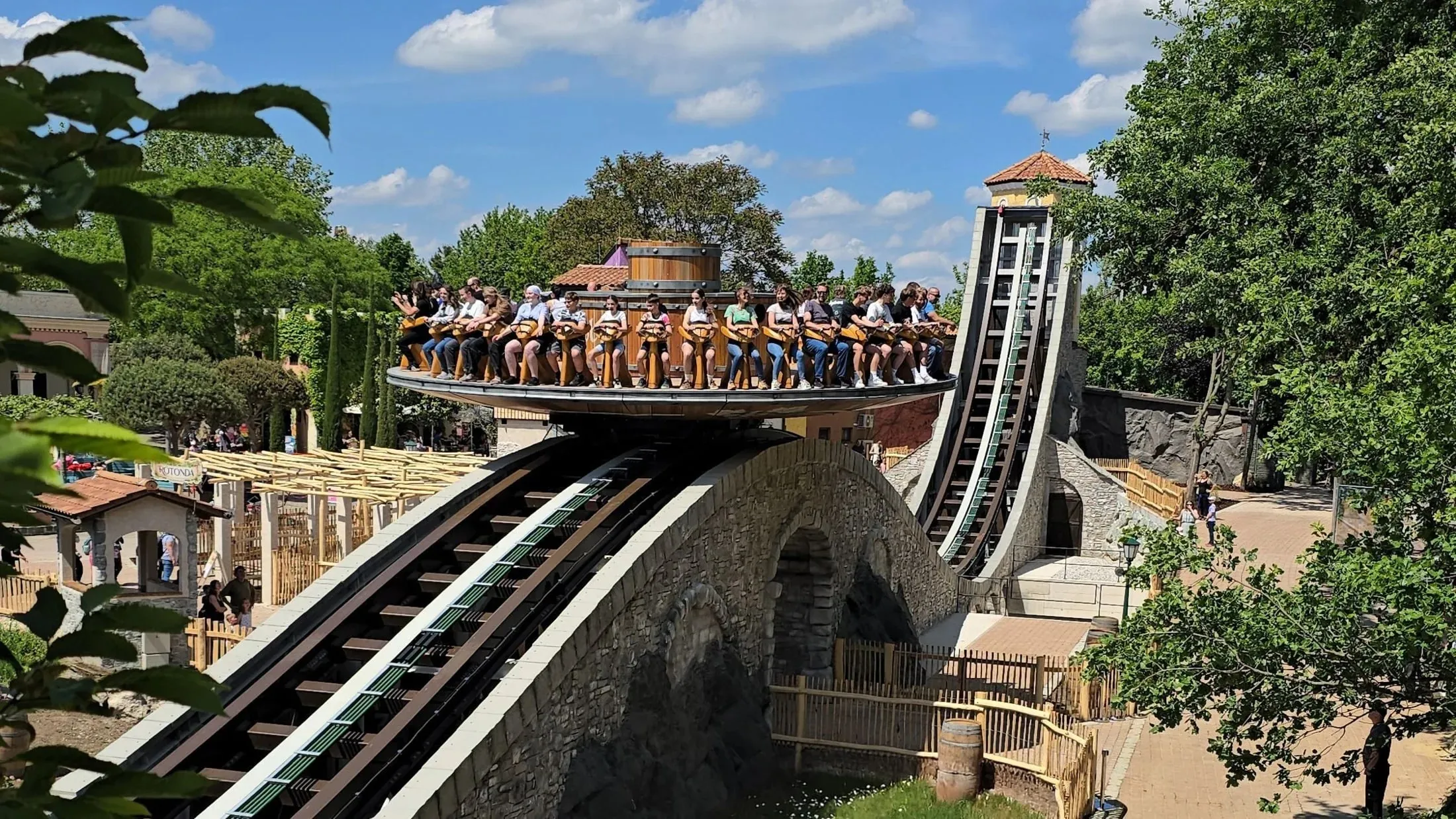 Achterbahn mit steinverkleideter Brücke und Felskulisse, Fahrgäste in der Scheitelposition